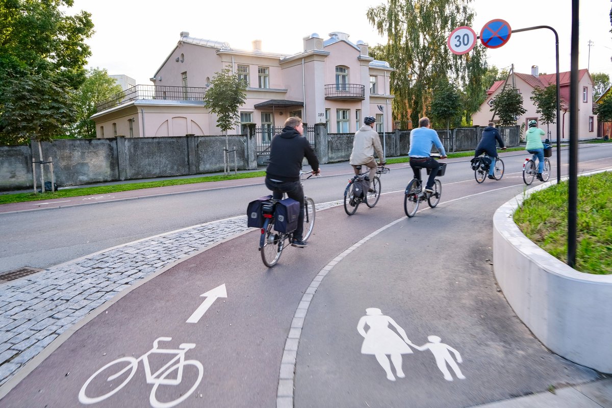 Cyclists in casual autumn attire ride along dedicated cycling infrastructure lined with buildings and trees on the streets of Tallinn, Estonia.