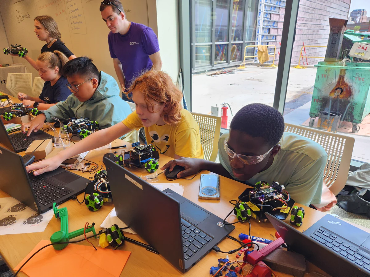 A group of individuals in a classroom are gathered around a table with laptops and small robotic devices. They appear to be engaged in a collaborative activity involving technology. A whiteboard in the background has writing and drawings. The environment is bright with natural light coming through large windows.