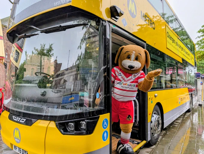 Boots the mascot dog in a red and white sports jersey waving from the doorway of a yellow Metro double-decker bus
