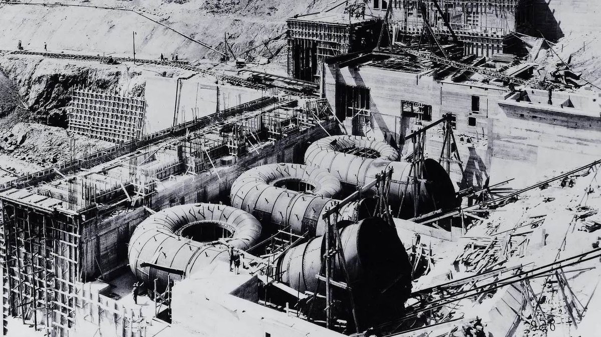 Historic black-and-white photo of hydroelectric dam construction showing large spiral turbine casings being installed. Workers visible among scaffolding, concrete forms, and excavated terrain.