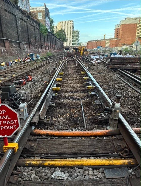 Railway track renewal work in an urban rail corridor, showing newly installed rails and track components in the foreground. Engineering materials, tools, and work equipment are positioned alongside the tracks, with brick retaining walls and city buildings visible in the background.