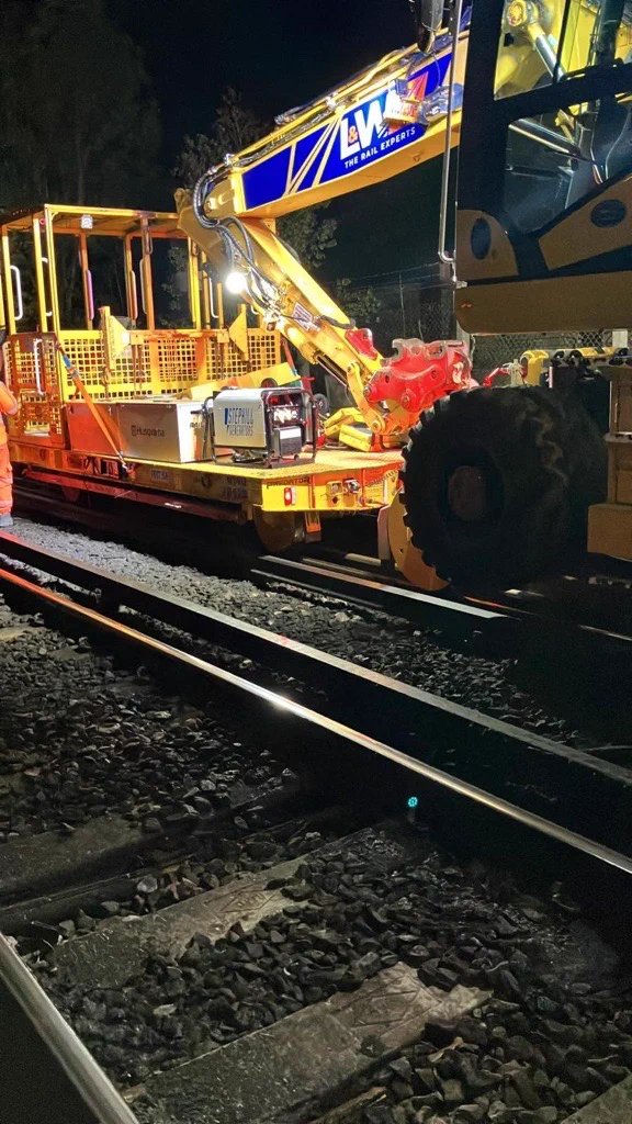 Rail-mounted excavator and engineering wagon operating on a railway line during overnight maintenance work. Bright work lights illuminate the machinery and track, with rail workers and equipment visible.