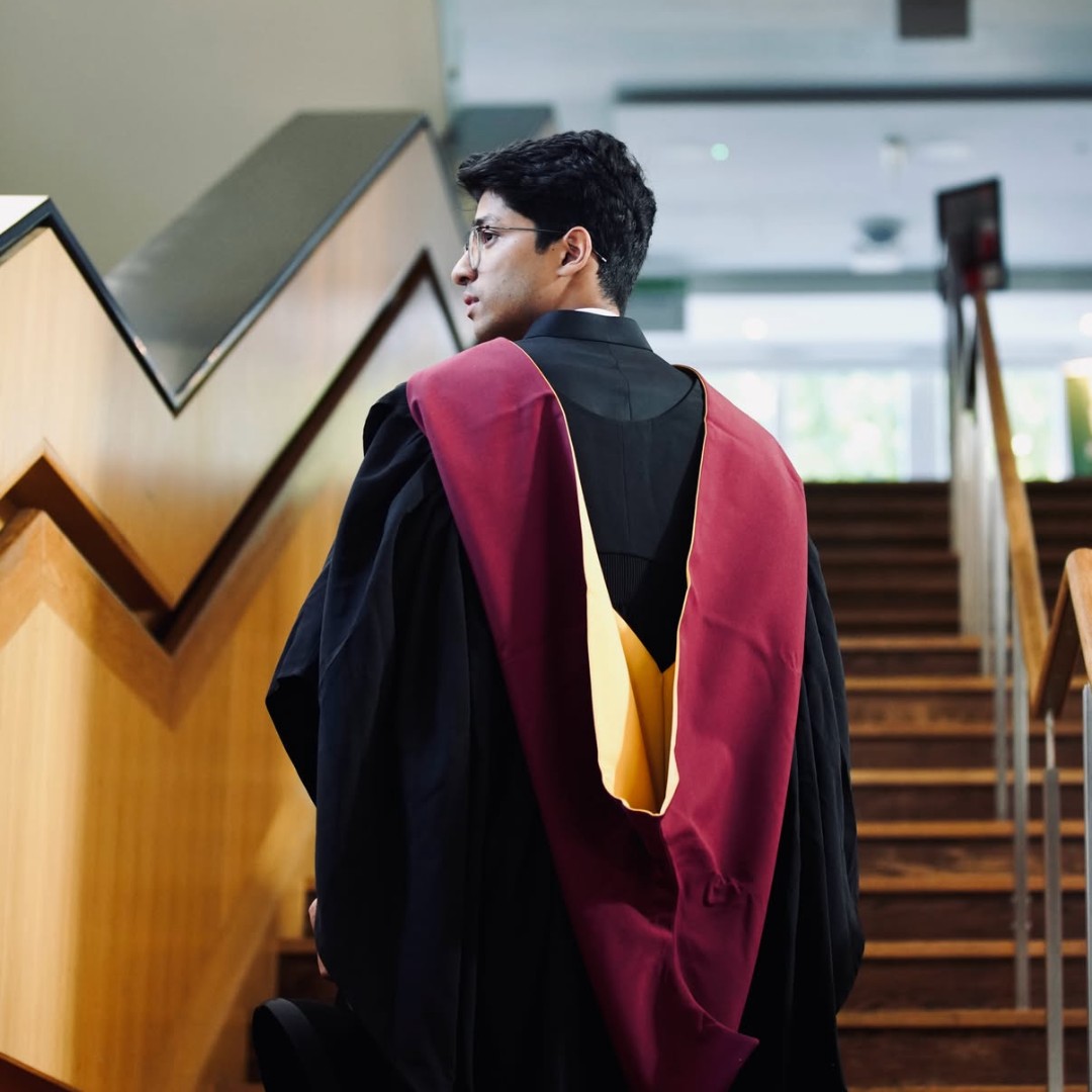 Young man in academic gown with maroon hood standing near wooden staircase indoors.