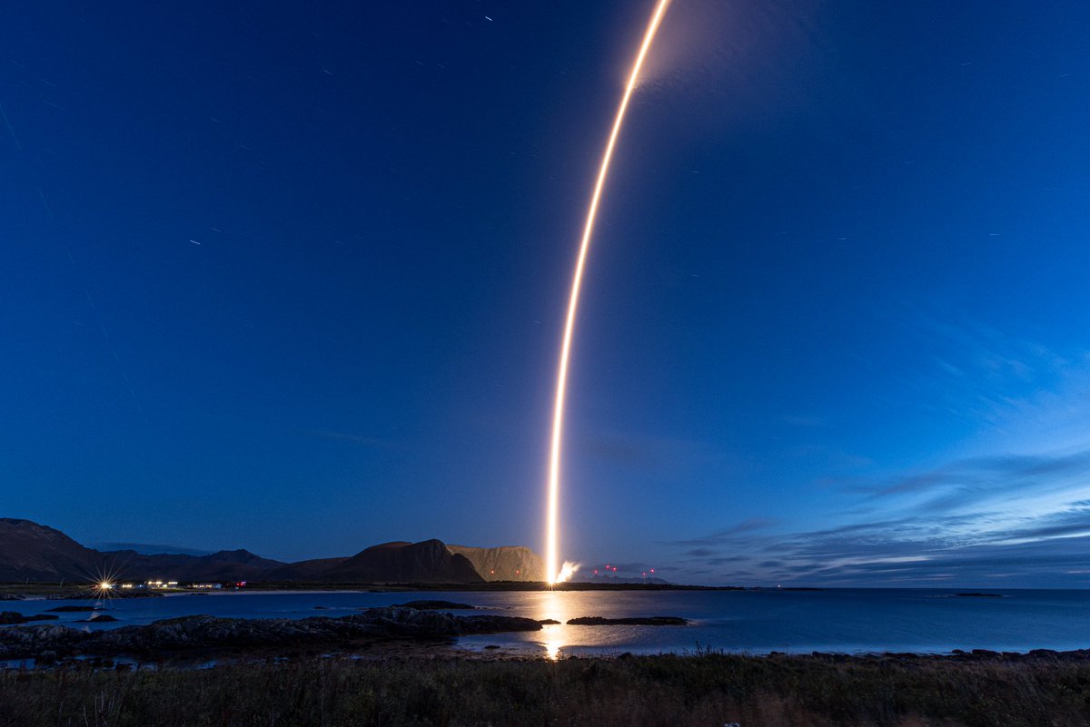Long-exposure photograph of a rocket launch at dusk. A bright arc of light rises from a coastal launch site across a deep blue sky, with mountains, calm water, and the rocket’s reflection visible below.