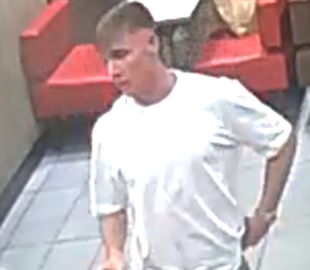 A grainy CCTV image of a young man with short hair wearing a white t-shirt, standing in front of red seating.