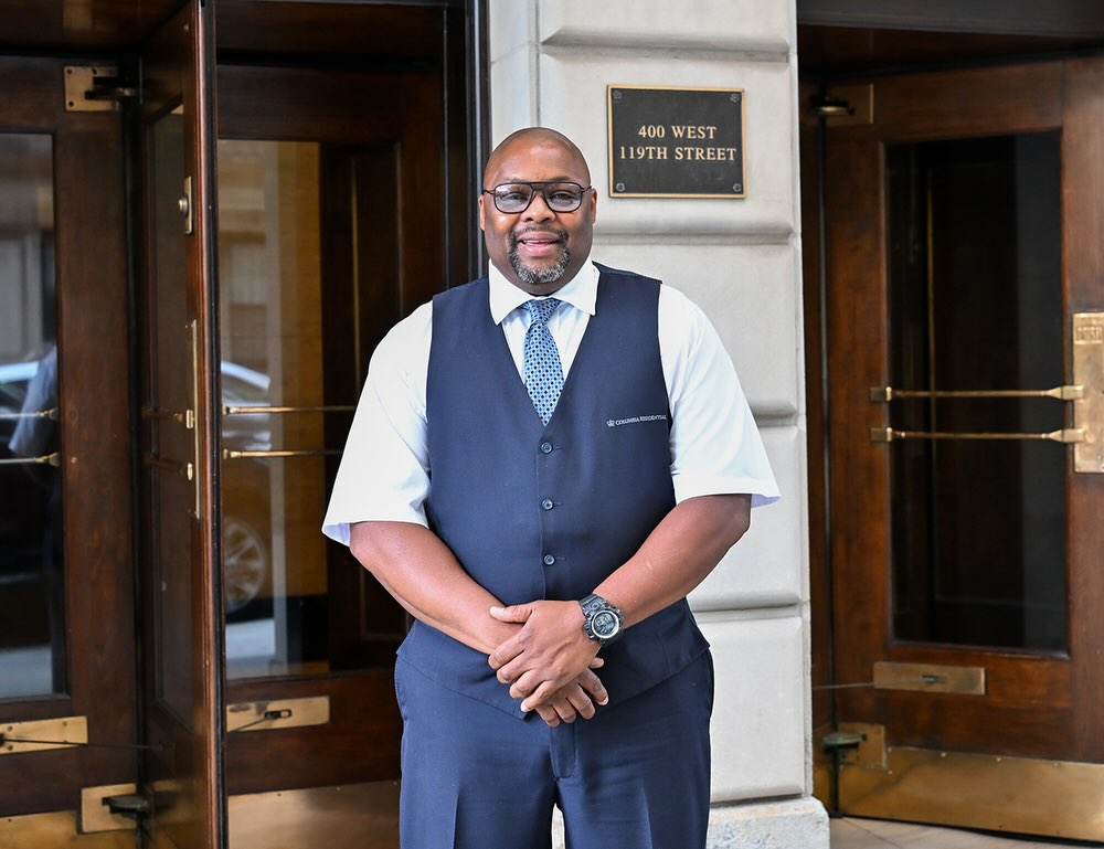 A Columbia Facilities doorman smiles and poses in front of a residential building at Columbia University.