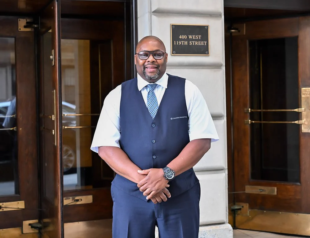 A Columbia Facilities doorman smiles and poses in front of a residential building at Columbia University.