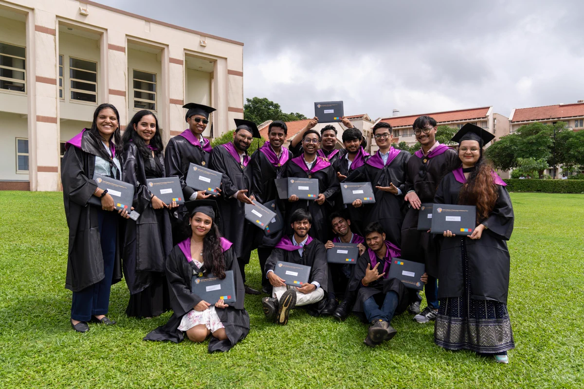 A diverse group of 18 smiling young adults, dressed in black graduation gowns with purple stoles and mortarboards, pose on green grass in front of a light-colored building. They hold up their gray certificates from 'Birla Institute of Technology & Science, Pilani'.