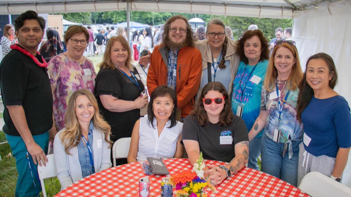 A group of people standing around a picnic table under a white tent. 