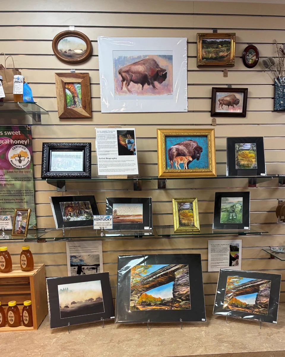A display wall features various framed paintings and photographs, including images of bison and landscapes. There are also bottles of honey on the left side, with a “Montana Sweet” label visible. The artworks are arranged on shelves and hooks against a slatted wall.
