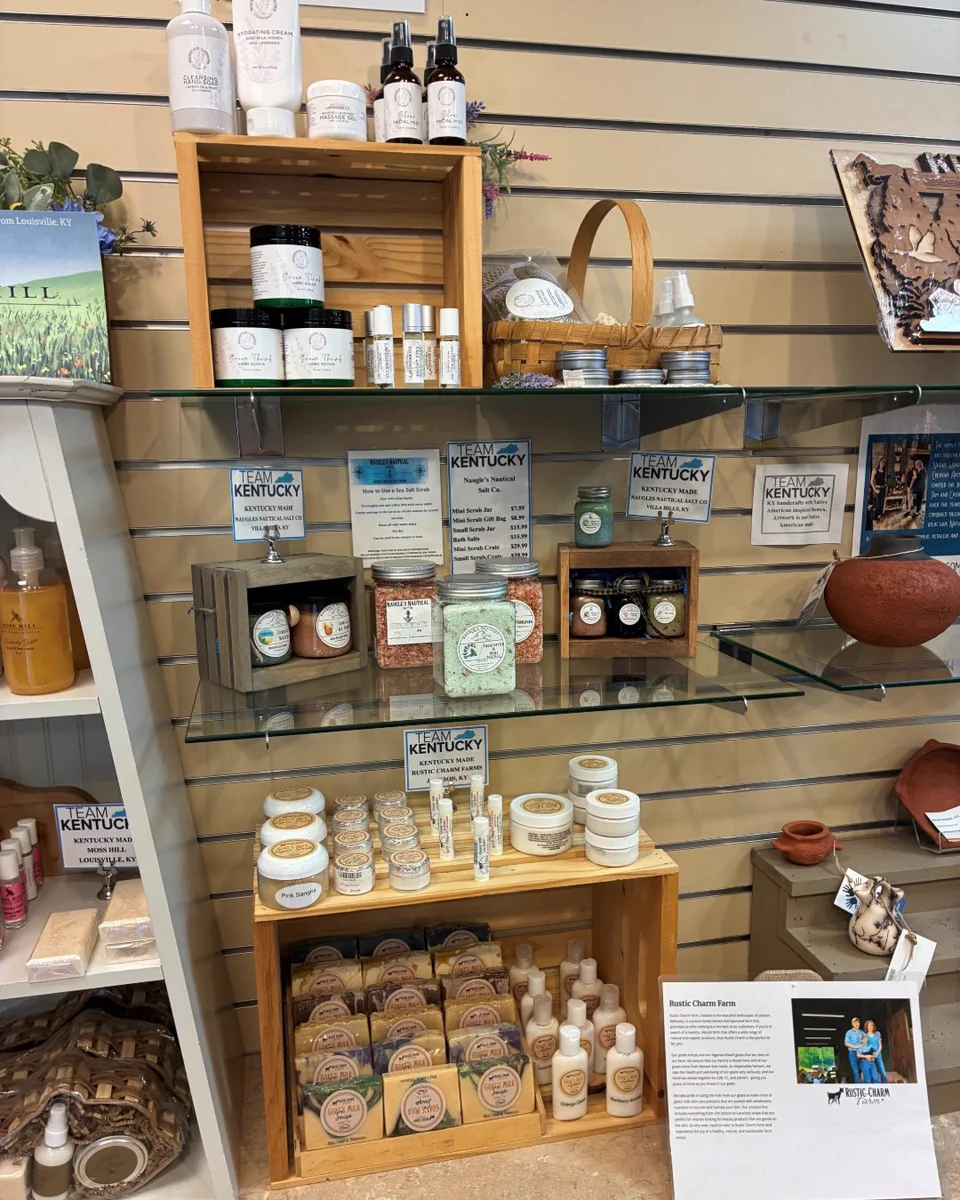 A display of various "Kentucky Proud" skincare products on wooden and glass shelves. Includes creams, lotions, oils, and soaps. Numerous bottles and jars are neatly arranged, accompanied by signage promoting local products. A basket and pottery piece are also visible nearby.