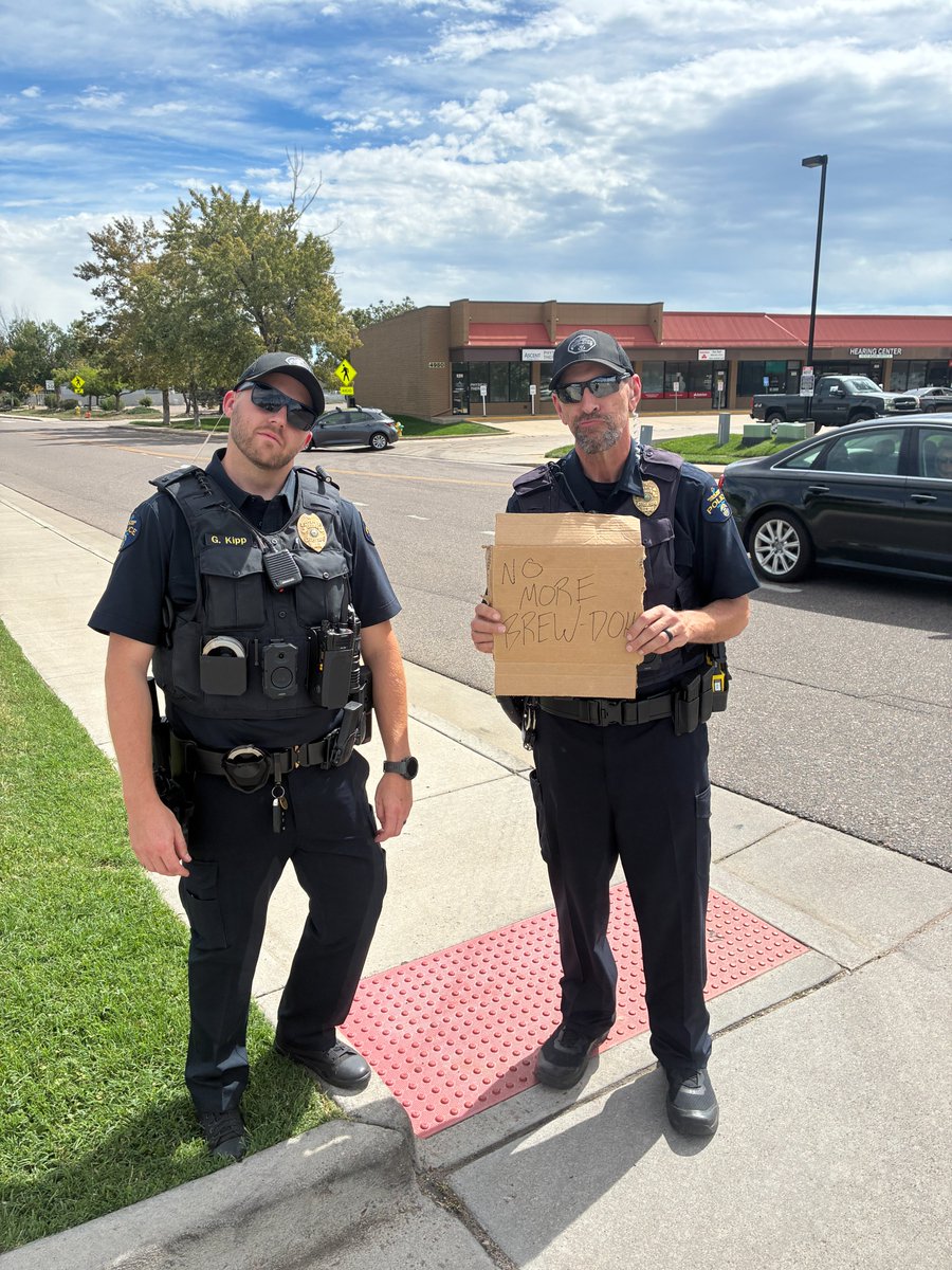 Two police officers stand outside a shopping center; one holds a cardboard sign reading “NO MORE BREW-DOH.”