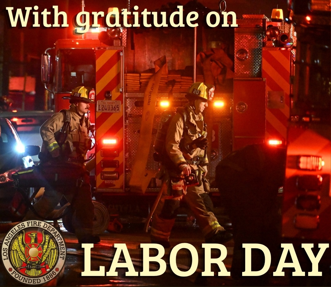 Two firefighters, in full turnout gear and carrying tools, walk with purpose past a red fire engine at night, glowing with emergency lights; text reads “With gratitude on Labor Day.”