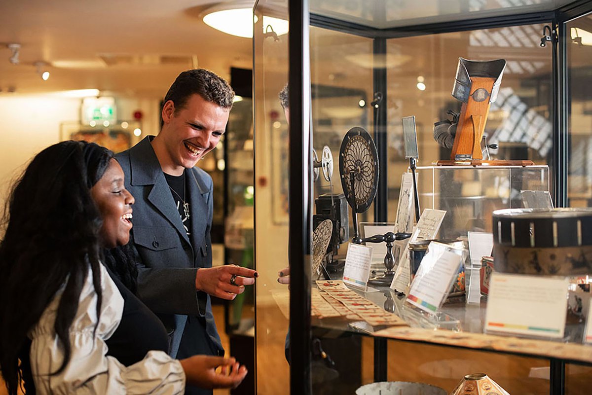 Two individuals are viewing exhibits in a museum display case at Bill Douglas Cinema Museum. They appear engaged and cheerful as they look at vintage film items with informational cards.