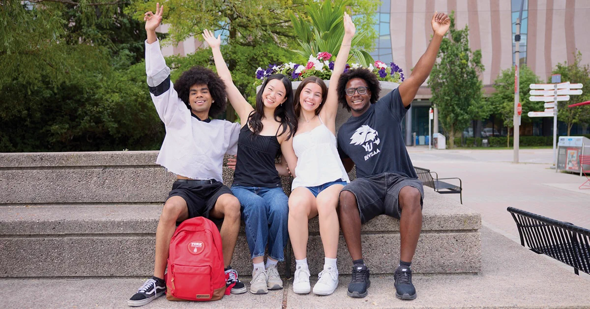 A group of four people sitting on outdoor steps, smiling with their arms raised. A bright red backpack is visible on the left. Flower arrangements and greenery are behind them, with a building in the background.