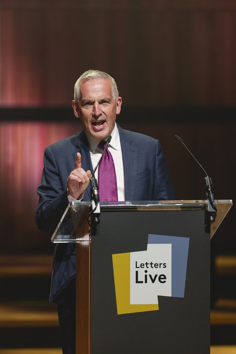 Chief Executive of the National Literacy Trust, Jonathan Douglas CBE, speaking at the 2026 Letters Live event. Jonathan is stood behind a lectern, is wearing a suit and has an animated expression.