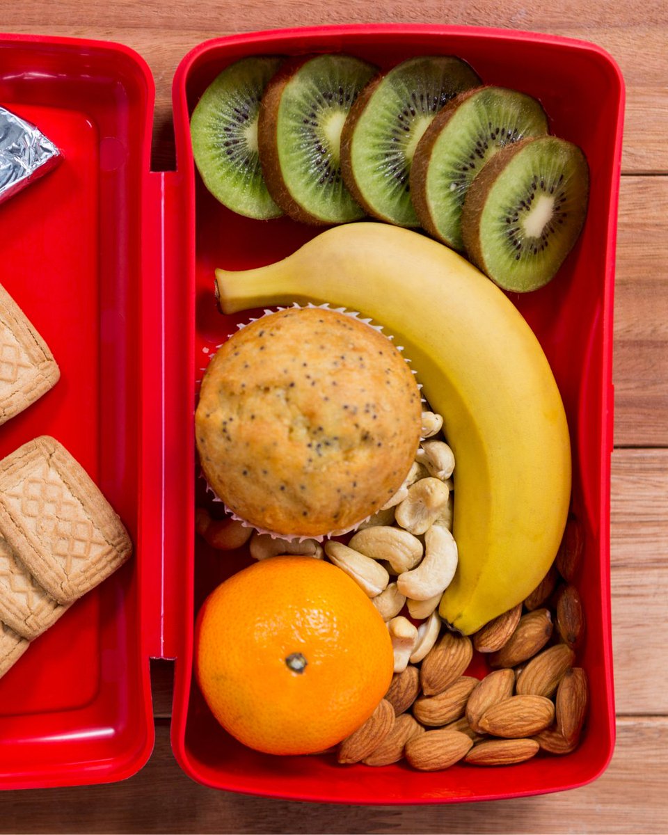 A red lunchbox containing sliced kiwi, a banana, a muffin, cashews, almonds, an Chilean mandarin.