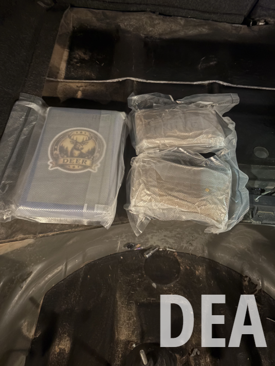 Two vacuum-sealed packages and a box with a deer logo placed inside a dark compartment, possibly a vehicle trunk. Suspected fentanyl powder.