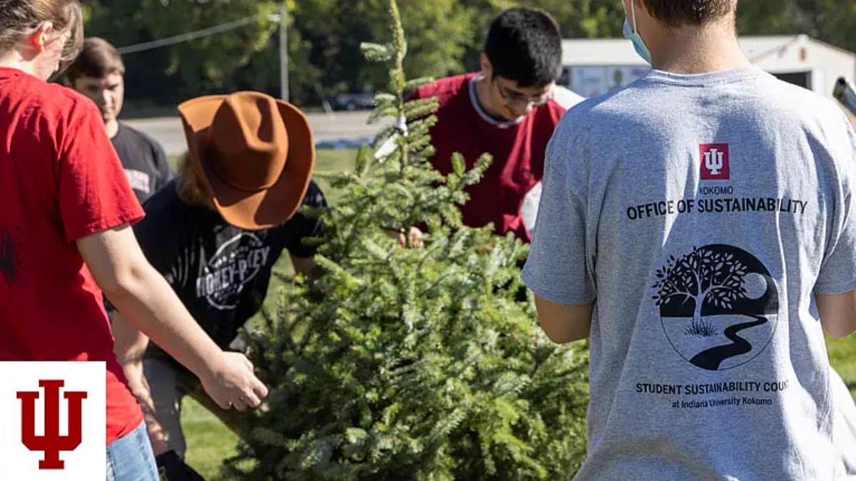 A group of people, some wearing Indiana University apparel, are gathered around a small evergreen tree outside. The Indiana University logo is visible in the corner of the image.