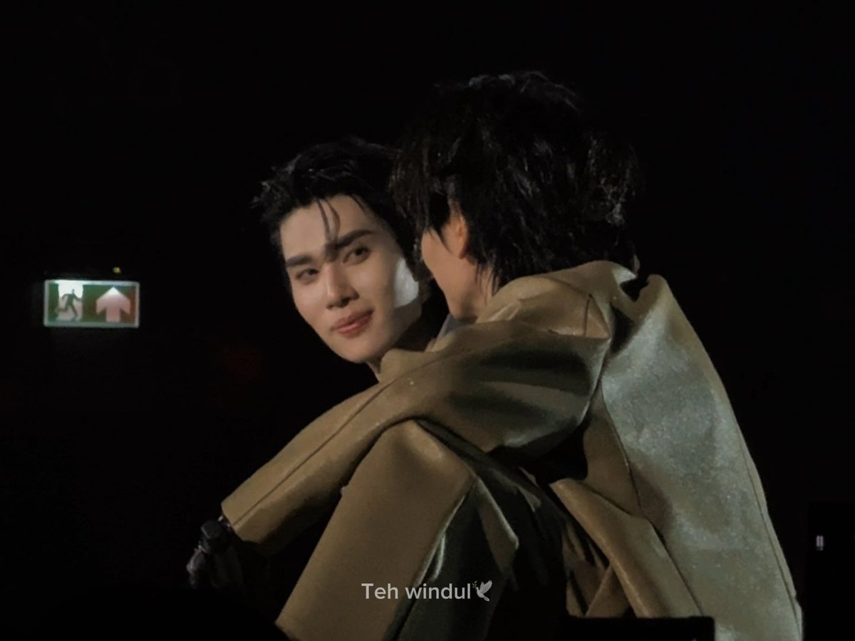 A K-pop performer (watermarked 'Teh windul') embraces another person on a dimly lit stage, looking over their shoulder at the camera near an exit sign.