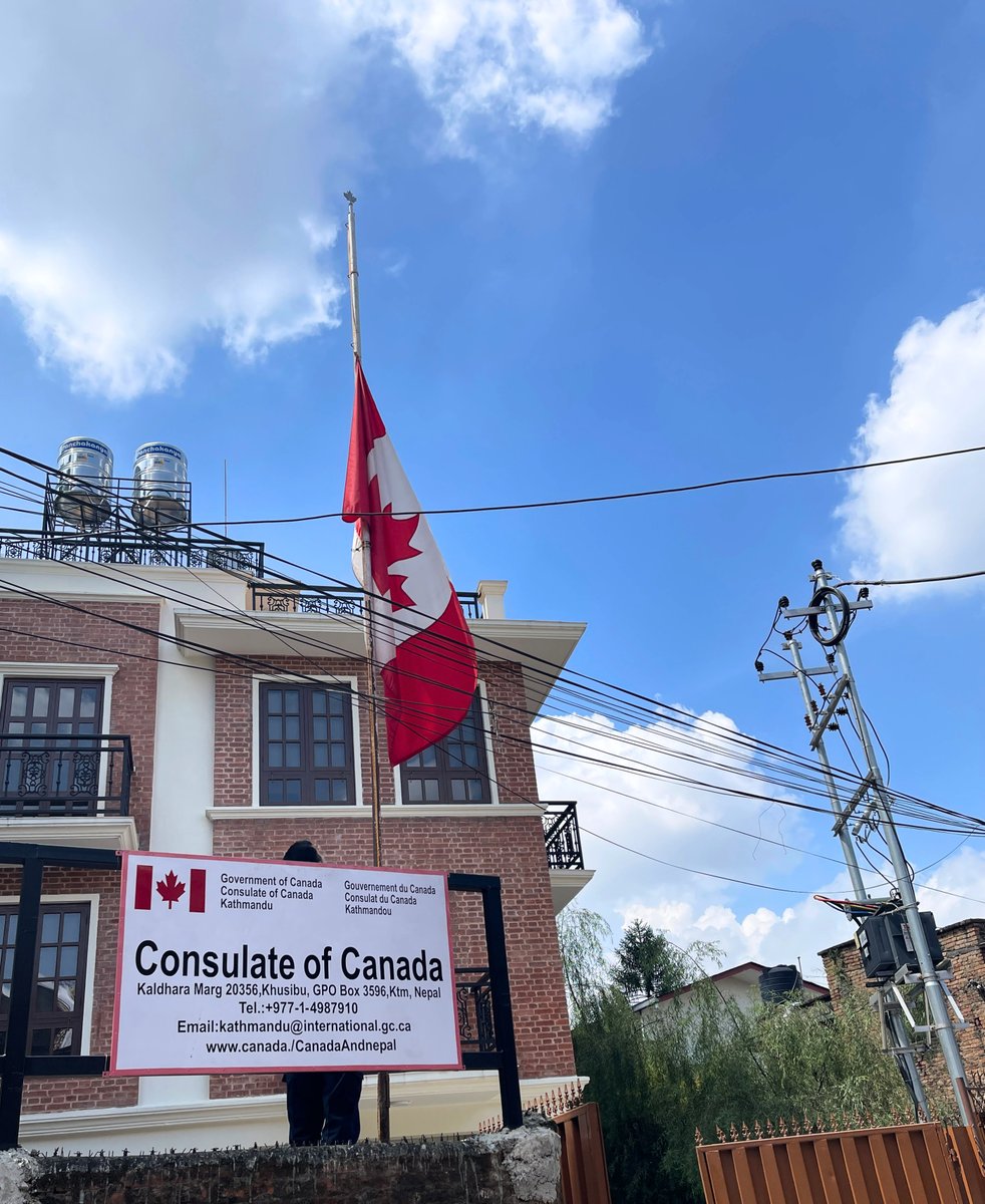 Canadian flag flying at half-mast in front of the Consulate of Canada in Kathmandu, Nepal. The consulate sign is visible in the foreground, with the building, utility lines, and a bright blue sky with scattered clouds in the background.