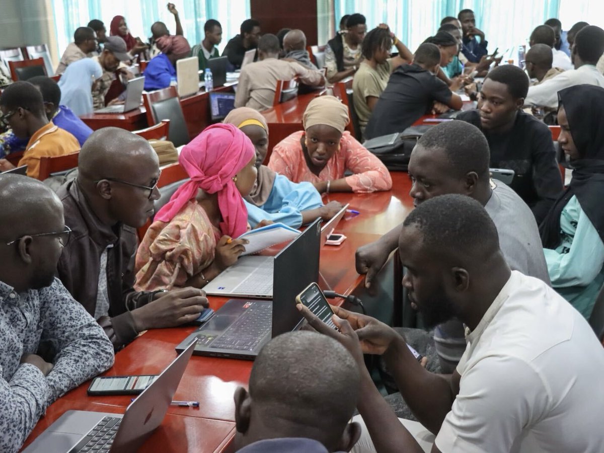 People sit in groups at tables, talking, looking at laptops, and looking at phones.