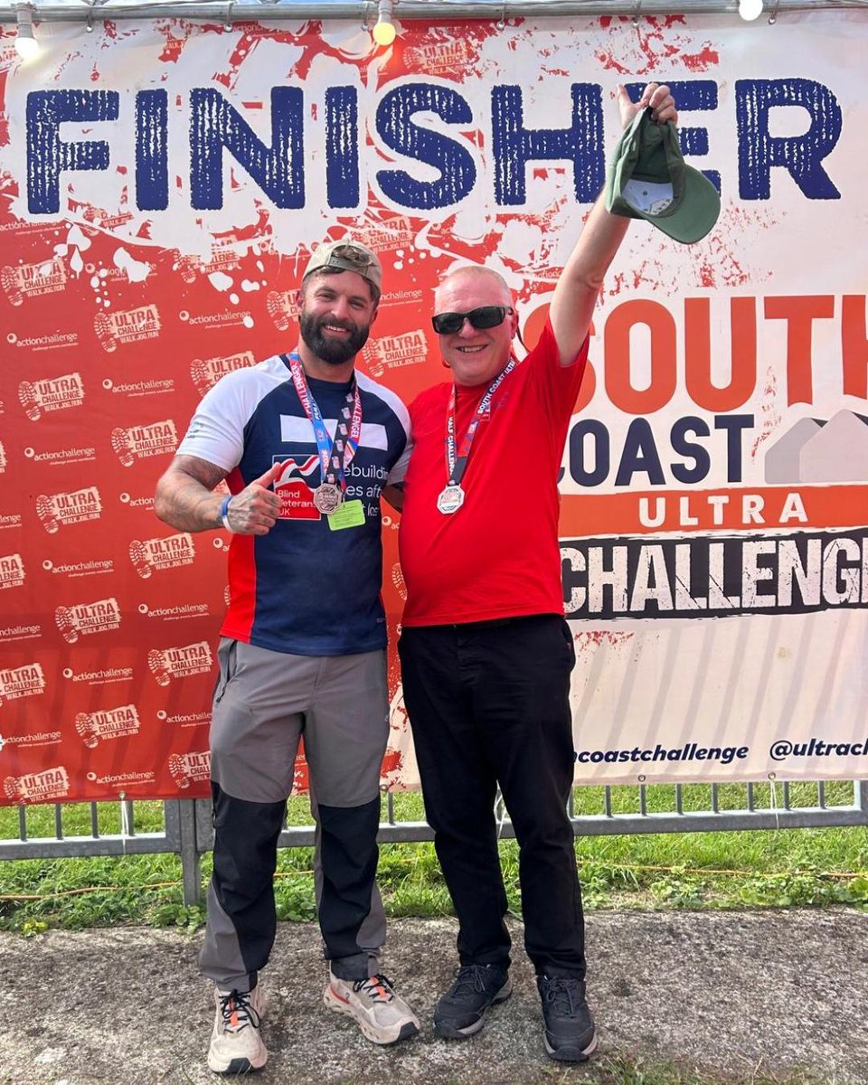 A Blind Veterans UK participant and guide stand together beneath the South Coast Ultra finish arch wearing finisher medals. The finish lane, event barriers and red challenge flags are visible behind them.