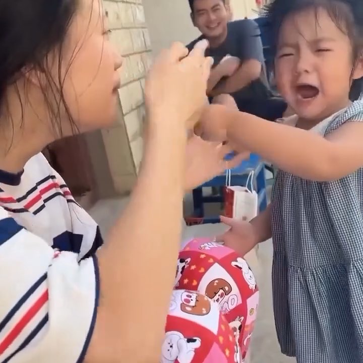 A candid family scene where a young girl in a checkered dress is laughing or crying while interacting with a woman, with a man sitting in the background and a pink patterned gift bag visible in the foreground.