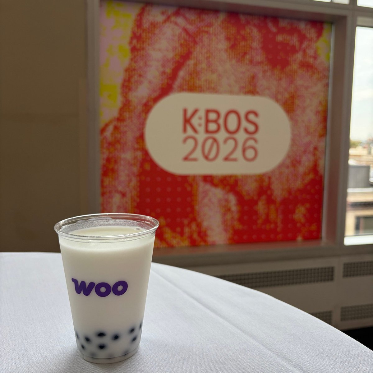 Bubble tea in a Woo-branded cup sits in front of a sign that says, "K:BOS 2026."