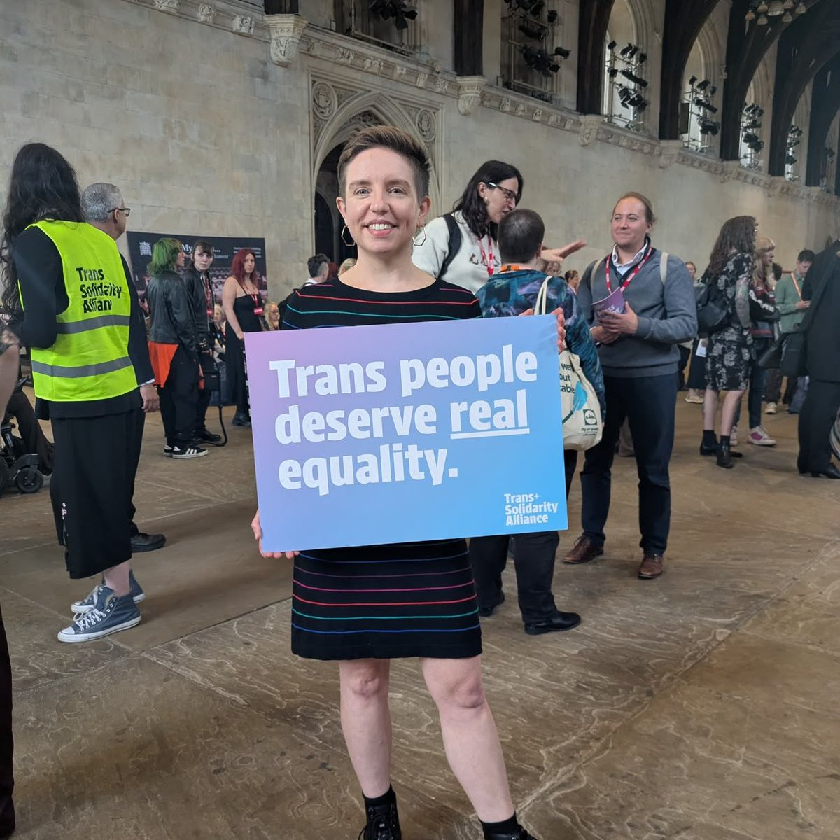 Carla stands in in an old part of Westminster in front of a crowd of people. She is holding a sign that reads "Trans people deserve real equality" (the word real is underlined).
