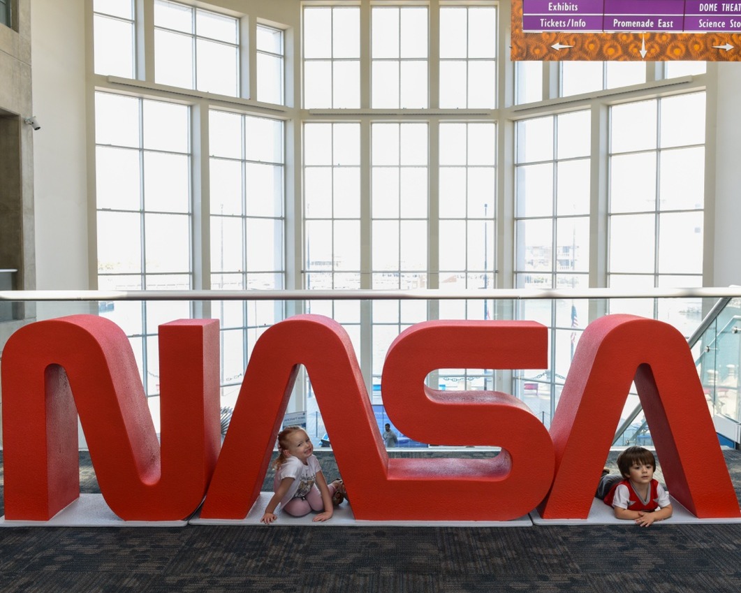 Two children lay underneath large letters spelling out "NASA" inside a large indoor museum space. Tall glass windows are visible behind them. Credit: NASA/Lily Hammel