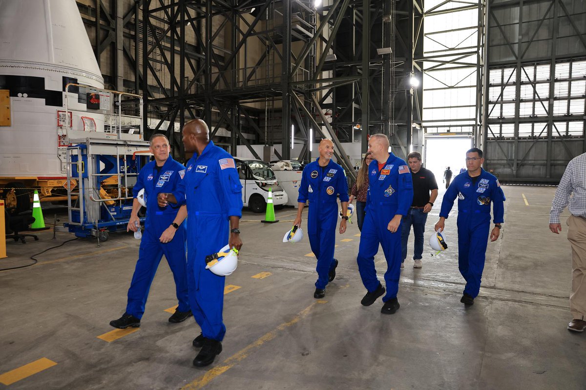 A group of people wearing blue flight suits walk together inside a large industrial facility, with tall metal structures and equipment in the background.
