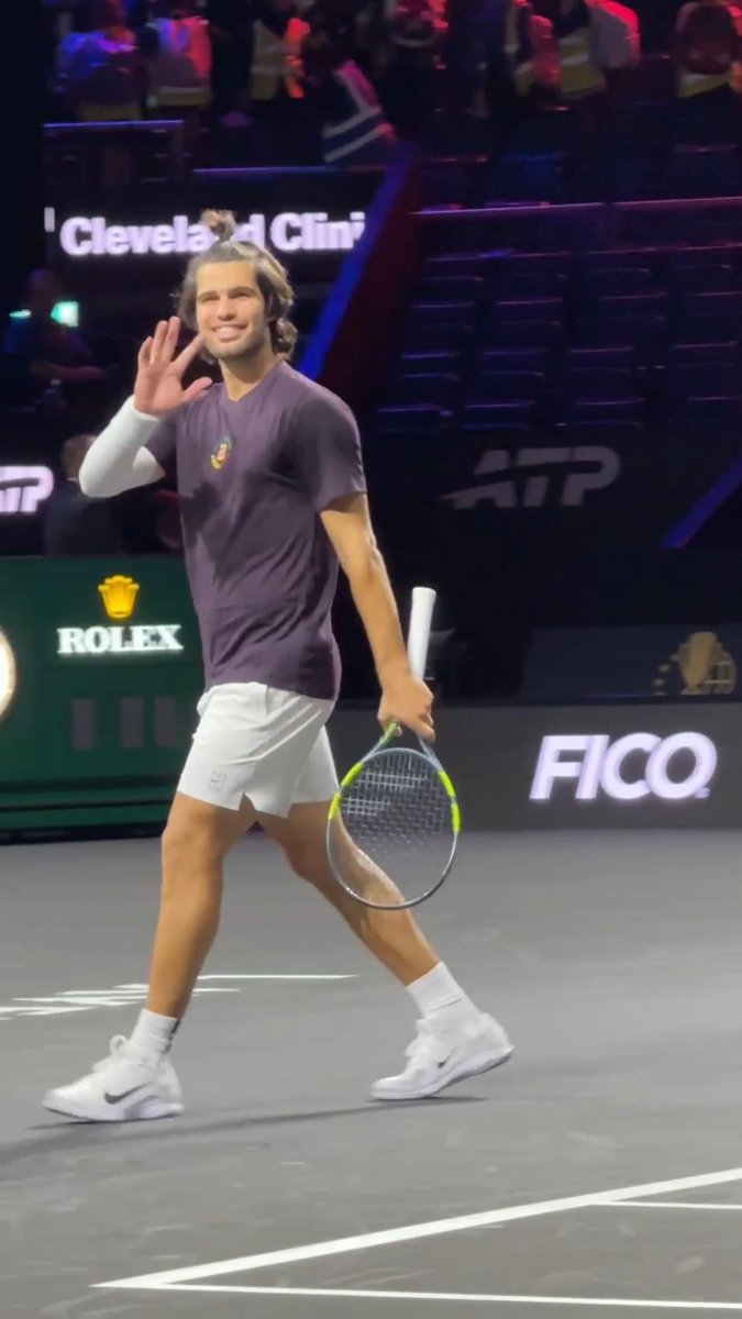 A tennis player in a purple shirt waves to the crowd while holding a racket on the court, with ATP, Rolex, and FICO sponsor logos visible in the background.