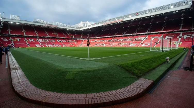 Empty red seats at Manchester United's Old Trafford stadium, with "STRETFORD END" visible in the stands, where small sections of the pitch are being sold to fans as souvenirs. The pristine green turf contrasts sharply with the vacant stadium, highlighting the high-quality grass being marketed in 7cm cubes.