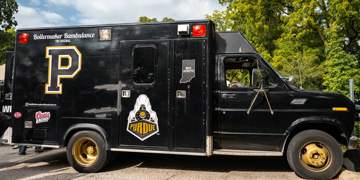 A black vehicle, modified like an ambulance, with a large "P" and Purdue University branding on the side. The doors display several logos and the "Boilermaker Bambulance" text. The vehicle is parked on a street with trees in the background.