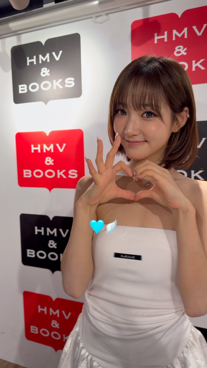 A young woman poses making a heart shape with her hands at an HMV & BOOKS event, wearing a white strapless dress with edited angel wings.