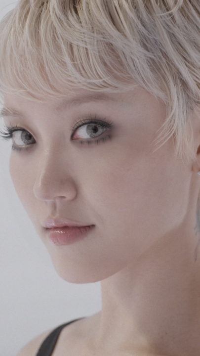 A close-up portrait of a model with a chic blonde pixie cut, dramatic lashes, and glossy lips, capturing a striking beauty look that complements the bold confidence referenced in the caption about her runway walk.