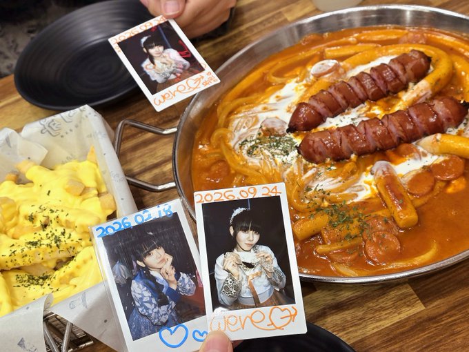 A meal of tteokbokki with sausages in a large pan alongside french fries, with several polaroid photos of a woman with dates '2026.09.24' and '2026.03.18' placed on the table.