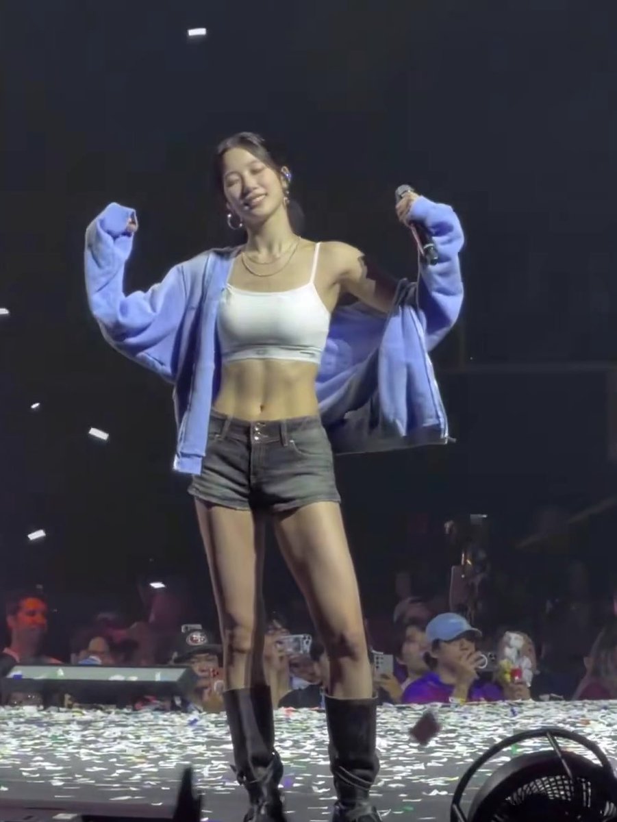 A woman in a white crop top, denim shorts, and blue cardigan poses with raised fists while holding a microphone on a concert stage with confetti scattered around.