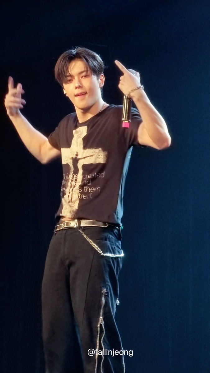 The image shows a male performer on stage wearing a dark graphic t-shirt featuring a distressed cross design with partially visible text reading "I...crucified...and...them...tre..." (likely "I am crucified with Christ"), paired with black pants and a chain belt, while pointing both index fingers upward. The watermark reads "@fallinjeong" and the caption's hashtags "#전이정 #leejeong #이정" indicate this is Lee Jeong (전 이정), suggesting fans are admiring and tagging the performer during a live concert or performance.
