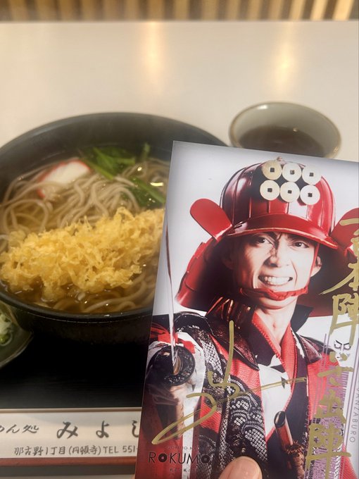 A hand holds up a signed promotional card featuring a man in samurai armor with a red helmet, held in front of a bowl of noodles at a restaurant identified as 'Miyamoto' (Karuizawa).