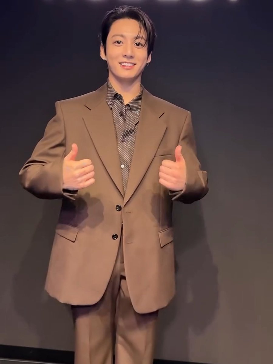 A man wearing a brown suit over a patterned shirt smiles brightly and gives two thumbs up to the camera.