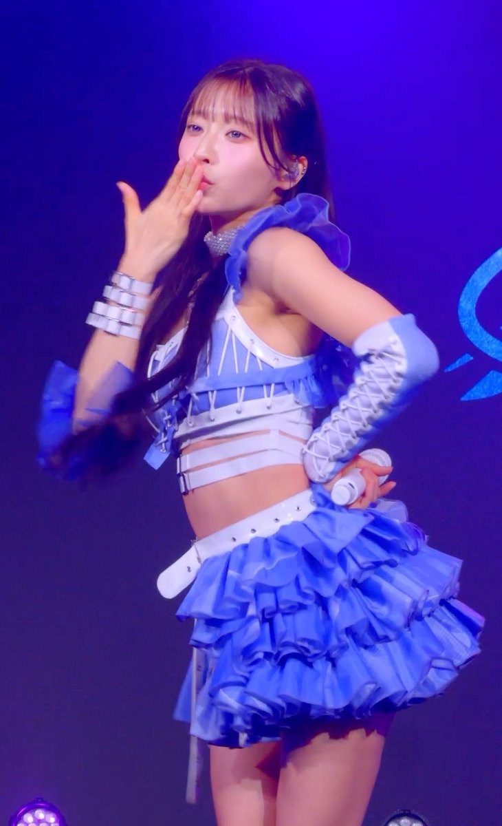 A photograph of a young woman performing on stage in a blue and white ruffled outfit, blowing a kiss towards the camera under purple lighting.
