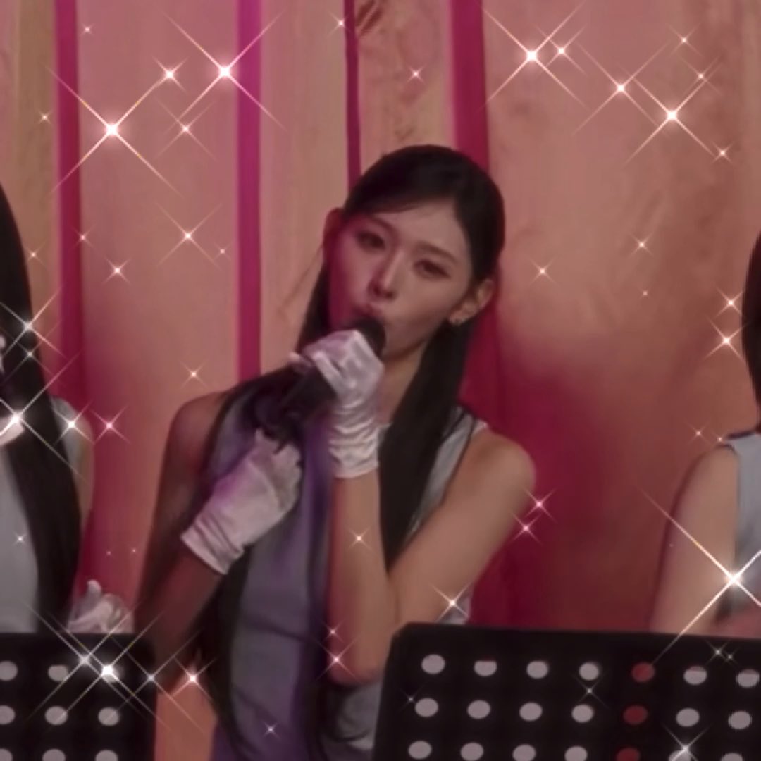 A screenshot with a heavy pink sparkle filter showing a woman with long dark hair wearing white gloves, holding a microphone and singing.