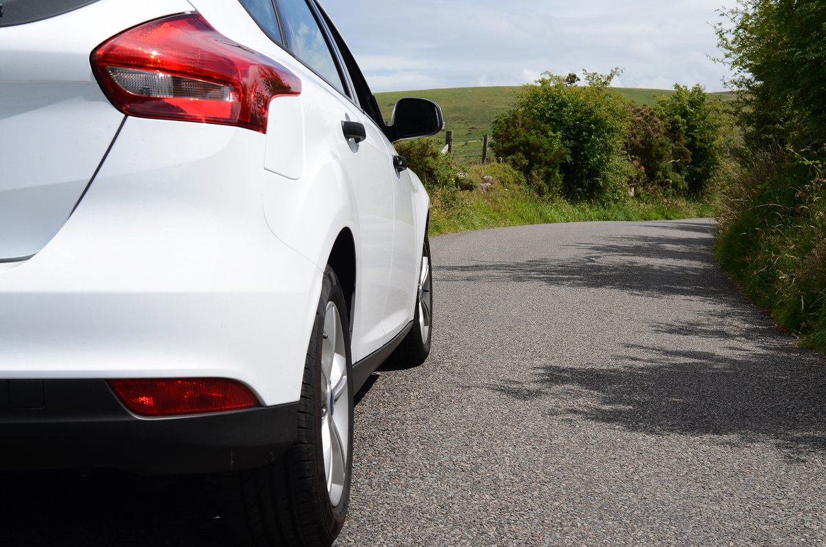 Rear side view of a white car parked on a quiet country road with green bushes and hills in the background.