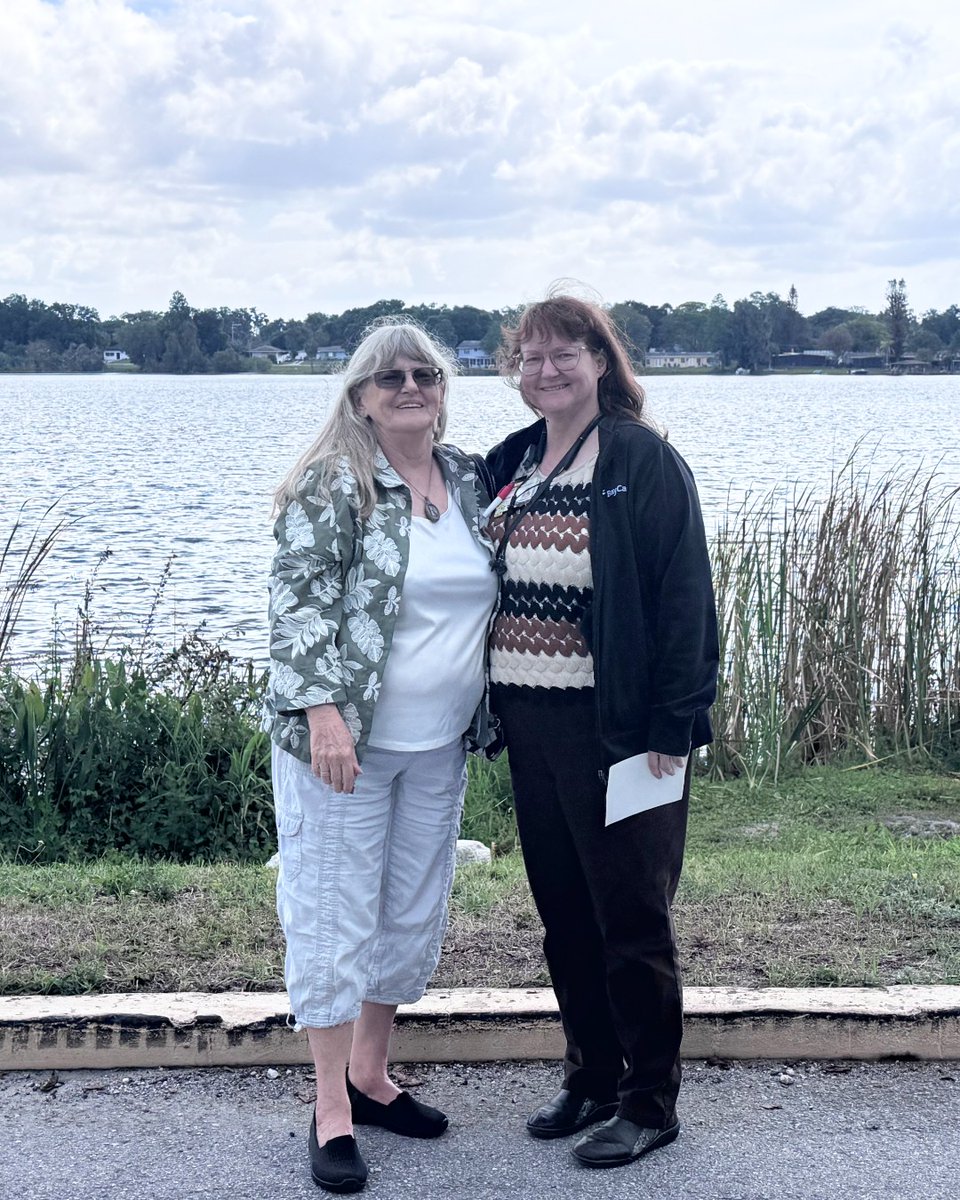 Two people are standing together in front of a lake. Both are smiling with greenery and water behind them under a partly cloudy sky.