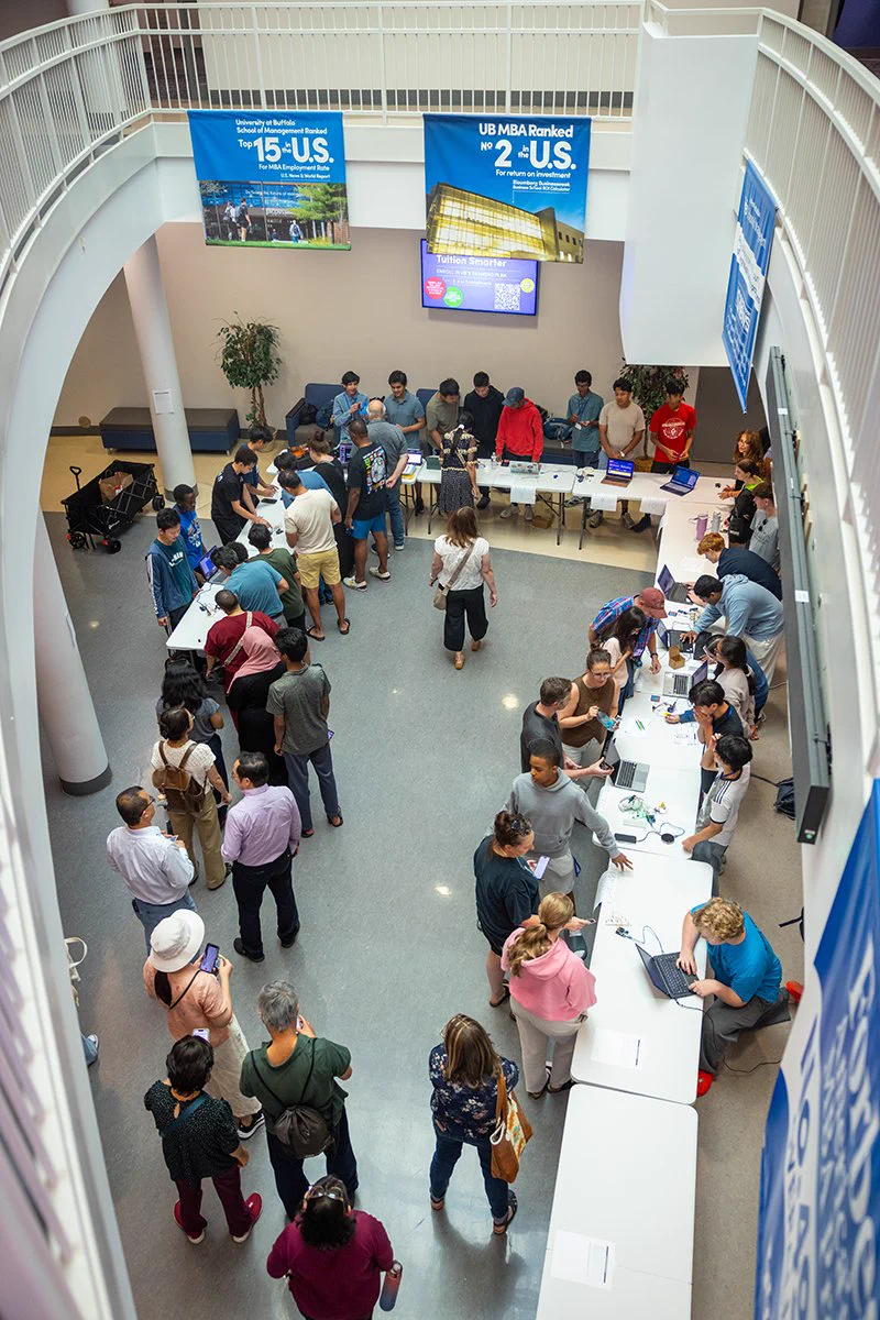 Overhead view of a crowded engineering showcase held in a large indoor atrium. Attendees visit project tables staffed by students demonstrating technology, engineering, and computing projects. Banners for the University at Buffalo School of Management hang above the event space while visitors move between exhibits and presentations.