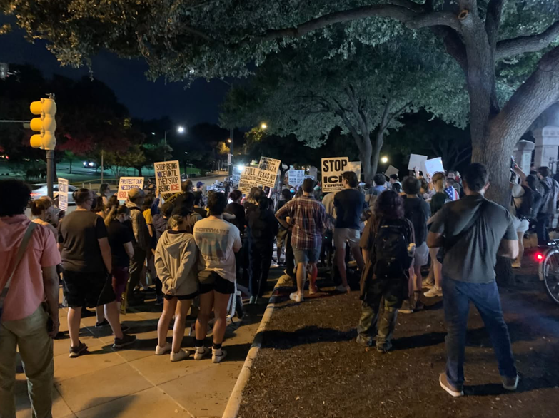 A nighttime protest or public demonstration on a sidewalk beneath large trees. A crowd faces forward, with several people holding signs. A traffic light and streetlights are visible nearby. The signs contain text.
