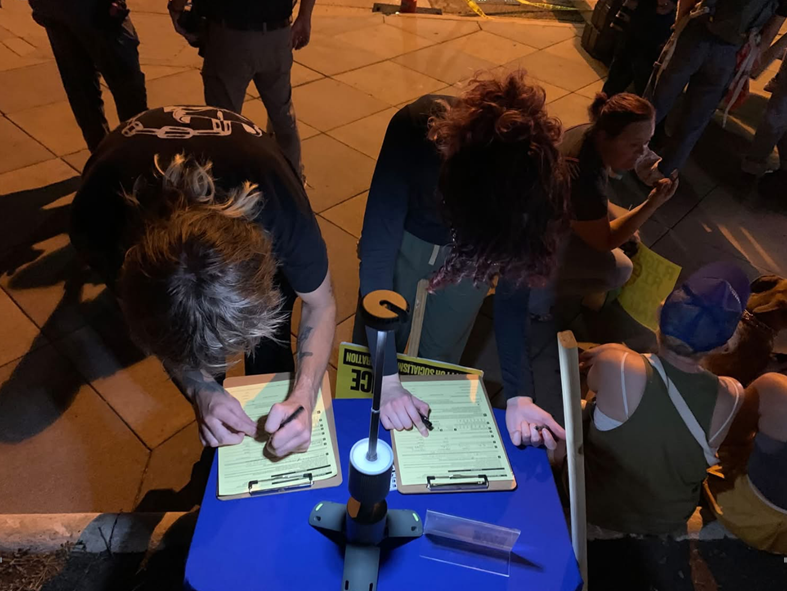 A nighttime street scene shows two people leaning over a blue table and filling out forms on clipboards, while other people sit or gather nearby. The scene is lit by warm artificial light, and text is visible on the forms and clothing.