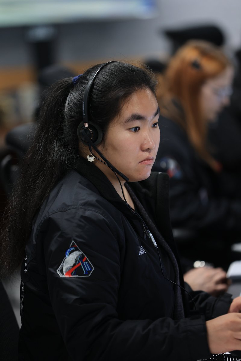 A woman wearing a black jacket with a NASA Artemis II logo on it, sits at a desk with a headset on surrounded by a group of others with headsets at NASA's Kennedy Space Center in Florida. Photo credit: NASA/Clayton Rougelot